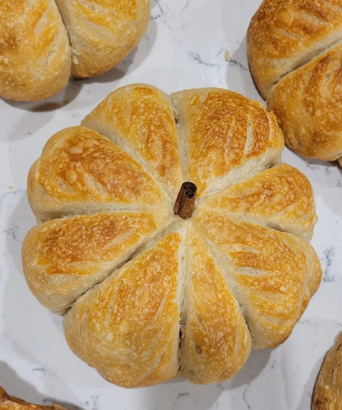 A golden loaf of bread shaped like a pumpkin, with a cinnamon stick for a stem, on a marble counter