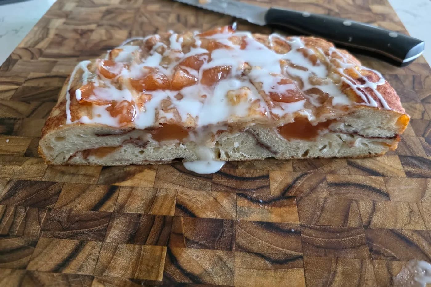 A fruit-topped pastry drizzled with white icing, cut to show the soft inside, on a wooden board with a bread knife
